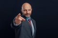 Portrait of bald man in business suit on dark background. Gentleman in his 40s with gray beard pointing finger in viewer direction Royalty Free Stock Photo