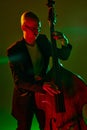 Portrait of bald musician in glasses plays double bass in red-green neon light against gradient studio background. Royalty Free Stock Photo