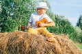 Portrait of barefoot boy in hat on haystack in field. Playing small guitar, ukulele. Light sunny day. Side view. Outdoor Royalty Free Stock Photo