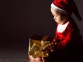 Portrait of barefooted infant baby boy in new year costume and red hat looking at his first new year gift golden box