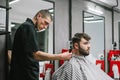 Portrait of bearded client cutting hair at hairdresser in men`s hairdresser, barber shaves neck trimmer with serious face. Male