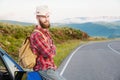 Portrait of a bearded happy serious traveler hipster with a backpack in a plaid shirt and a hat next to an unknown car