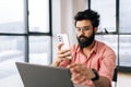 Portrait of bearded Indian businessman in glasses holding mobile phone texting online message sitting at office desk by Royalty Free Stock Photo