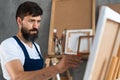 portrait of a bearded male artist in a dirty apron stands in an art studio, holding a brush in his hands and paints a Royalty Free Stock Photo