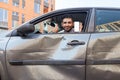 Portrait of bearded man in crashed car looking at camera and smiling, showing thumb up and smart phone with blank screen,