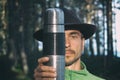 Portrait of bearded millennial man in felt hat holding stainless thermos near his face on woods background. Tourist adventurer on