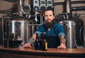 Portrait of a bearded tattooed hipster male in a jeans shirt and apron working in a brewery factory, standing behind a Royalty Free Stock Photo