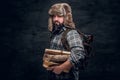 Portrait of a bearded woodcutter with a backpack dressed in a plaid shirt and trapper hat holding firewood.