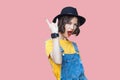 Portrait of beautiful amazed young woman in yellow t-shirt, blue denim overalls with makeup and black hat standing with rock horns Royalty Free Stock Photo