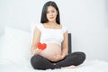 Portrait of beautiful Asian pregnant woman sit on bed and hold red heart icon also smiling and look to camera. Concept of good Royalty Free Stock Photo