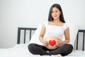 Portrait of beautiful Asian pregnant woman sit on bed and hold red heart icon also smiling and look to camera. Concept of good Royalty Free Stock Photo