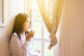 Portrait of beautiful asian woman is holding a cup of coffee and looking something on window at home in the morning,Happy and smil