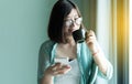 Portrait of beautiful asian women is holding a cup of coffee and using mobile phone on window at home in the morning,Happy and smi