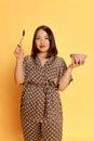 Portrait of beautiful, asian, young girl in silk homewear, pajamas standing with bowl and spoon against yellow studio Royalty Free Stock Photo