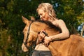 Portrait of a beautiful blond woman with her arms around the neck of her yellow foal. In autumn sunlight, trees in background