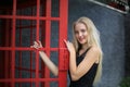 Portrait of Beautiful blonde hair girl on black dress standing in red phone booth against black wall as portrait fashion pose outd Royalty Free Stock Photo