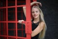 Portrait of Beautiful blonde hair girl on black dress standing in red phone booth against black wall as portrait fashion pose outd Royalty Free Stock Photo