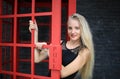 Portrait of Beautiful blonde hair girl on black dress standing in red phone booth against black wall as portrait fashion pose outd Royalty Free Stock Photo