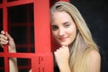 Portrait of Beautiful blonde hair girl on black dress standing in red phone booth against black wall as portrait fashion pose outd Royalty Free Stock Photo