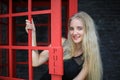 Portrait of Beautiful blonde hair girl on black dress standing in red phone booth against black wall as portrait fashion pose outd Royalty Free Stock Photo