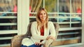 Portrait of beautiful blonde smiling young woman sitting on chair waiting in city cafe Royalty Free Stock Photo