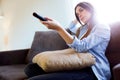 Beautiful bored young woman watching TV and holding remote control at home.