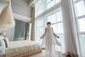 Portrait of beautiful bride in white silk dressing gown with modern hairstyle and long veil standing near window in bedroom.