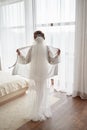 Portrait of beautiful bride in white silk robe with curly hairstyle and long veil standing near window in bedroom, copy space.