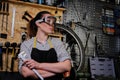 Portrait of a beautiful brunette female wearing working clothes, apron and goggles, standing in a workshop. Royalty Free Stock Photo