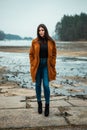 Portrait of beautiful brunette girl woman in orange fashion coat in season with frozen lake on background. Female model in jeans Royalty Free Stock Photo