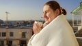 Portrait of beautiful brunette woman covered in blanket drinking coffee and looking on the city skyline Royalty Free Stock Photo