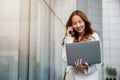 Asian businesswoman working on laptop and talking on cell phone at front building near office Royalty Free Stock Photo