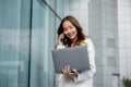 Asian businesswoman working on laptop and talking on cell phone at front building near office Royalty Free Stock Photo