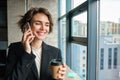 Portrait of beautiful businesswoman in suit, drinking coffee in office and talking on mobile phone, smiling happily Royalty Free Stock Photo