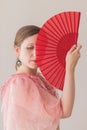 Portrait of a beautiful caucasian young woman in a pink dress covering half of her face with a red fan Royalty Free Stock Photo