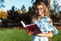 Portrait of a beautiful coed girl listening to music on headphones, outdoors, wearing glasses, holding a book and reading it Royalty Free Stock Photo