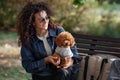 Portrait of beautiful curly haired woman with tiny poodle smiling, sits outdoors in park. Pet with its owner in nature