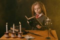 Little witch dressed in dark clothes sitting at the table against a black smoky background. There are magic wand, books Royalty Free Stock Photo