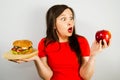 Portrait of a beautiful fat girl in surprise and holding a red apple and a hamburger, on gray background