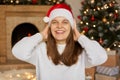 Portrait of beautiful female model wearing santa hat and white jumper, keeping hands on her head, posing in living room with Royalty Free Stock Photo