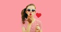 Portrait of beautiful girl blowing her lips with red lipstick holding red sweet heart shaped lollipop on pink background Royalty Free Stock Photo