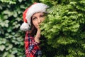 Portrait of a beautiful girl with a dreaming, piercing look in a santa claus red hat looks away through green branches of juniper.