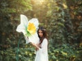 Portrait of a beautiful girl elf with long hair stands in a fantasy forest and hugs a huge daffodil flower, green trees Royalty Free Stock Photo