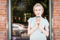 Portrait beautiful girl with long blonde hair holds a coffee cup stands by the window of a street city cafe, looks to camera Royalty Free Stock Photo