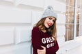 Portrait beautiful girl with long hair in marsala sweater on grey wall background outside. She wears knitted hat, red