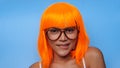 Portrait of beautiful happy Asian woman in bright orange wig and glasses posing over blue Royalty Free Stock Photo