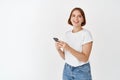 Portrait of beautiful happy girl holding smartphone screen and smiling, listening music in wireless headphones, standing Royalty Free Stock Photo