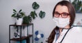 Portrait of beautiful happy young Caucasian female doctor wearing lab coat, face mask posing at clinic office workplace. Royalty Free Stock Photo