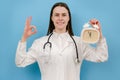 Portrait of beautiful happy young woman doctor holding alarm clock, showing ok sign, wears white uniform and stethoscope, looking Royalty Free Stock Photo