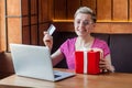 Portrait of beautiful happy young woman with short hair in pink t-shirt and eyeglasses is sitting in cafe, holding credit card and Royalty Free Stock Photo